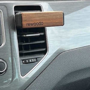 Könnte beinhalten: Ein rechteckiger Auto-Luftauslassdiffusor aus Holz mit dem Aufdruck "rewoodo". Der Diffusor besteht aus dunklem Holz und hat einen schwarzen, gummierten Rand. Er wird in eine Lüftungsöffnung des Autos eingesetzt.