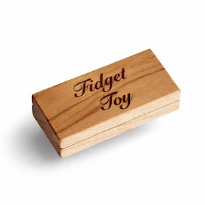 Op de afbeelding: Een rechthoekige houten doos met de tekst "Fidget Toy" gegraveerd op de bovenkant. De doos is gemaakt van lichtbruin hout met zichtbare houtnerf. De doos is gesloten, wat duidt op een kleine container.