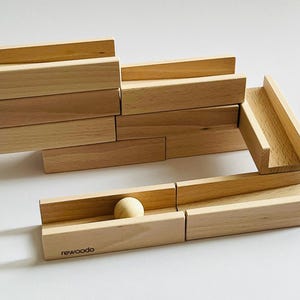 Könnte beinhalten: Ein hölzernes Murmelbahnspielzeug mit einer Kugel in einer Bahn. Das Set enthält verschiedene hellbraune Holzblöcke und Bahnteile, die so angeordnet sind, dass sie einen Weg für die Kugel bilden. Die Kugel ist hellbeige. Der Markenname "rewoodo" ist auf einem der Blöcke aufgedruckt.