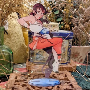 May include: A clear acrylic figure of a woman in orange pants and a white top, holding a blue and white device. The figure is on a blue base, set on a carved wooden stand. The figure is a character from the video game Portal.