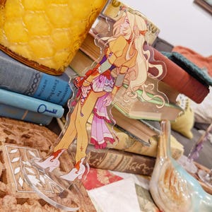 May include: A clear acrylic standee featuring a cartoon character with long blonde hair, wearing a yellow top, pink skirt, and white boots with red accents. The standee is placed in front of a stack of old books and a jar of honey.