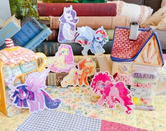 May include: A collection of six colorful pony stickers featuring various characters, arranged in front of a stack of vintage books and a ceramic tea room teapot. The stickers display vibrant illustrations of winged and earth ponies in dynamic poses.