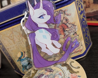 May include: A clear acrylic charm featuring a white unicorn character with a purple mane and tail, blue eyes, and a cutie mark of three blue diamonds. The charm has a subtle glitter finish and is attached to a white plastic ring connector.