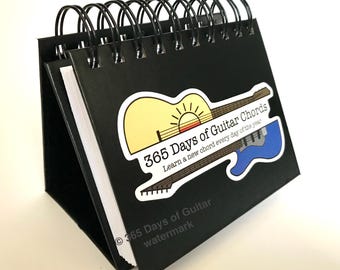 Könnte beinhalten: Ein schwarzes Spiralbuch mit einem Aufkleber auf der Vorderseite. Der Aufkleber zeigt einen gelben und orangen Sonnenuntergang mit einer Gitarrensilhouette und dem Text "365 Days of Guitar Chords Learn a new chord every day of the year". Am unteren Rand des Buches befindet sich ein Wasserzeichen mit dem Text "365 Days of Guitar watermark".