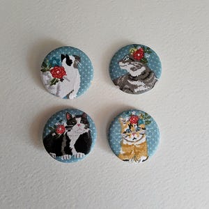 May include: Four round fabric buttons with cat and dog illustrations, each adorned with floral accents. The backgrounds are light blue with white polka dots. The animals are in various colors, including black, white, gray, and orange.