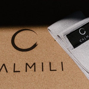 K&ouml;nnte beinhalten: Ein graues Stoff-Widerstandsband mit dem Wort "CALMILI" und einem Kreislogo, das auf einem schwarzen Etikett gedruckt ist. Das Band liegt auf einer Korkoberfl&auml;che mit dem Wort "CALMILI" in Schwarz gedruckt.