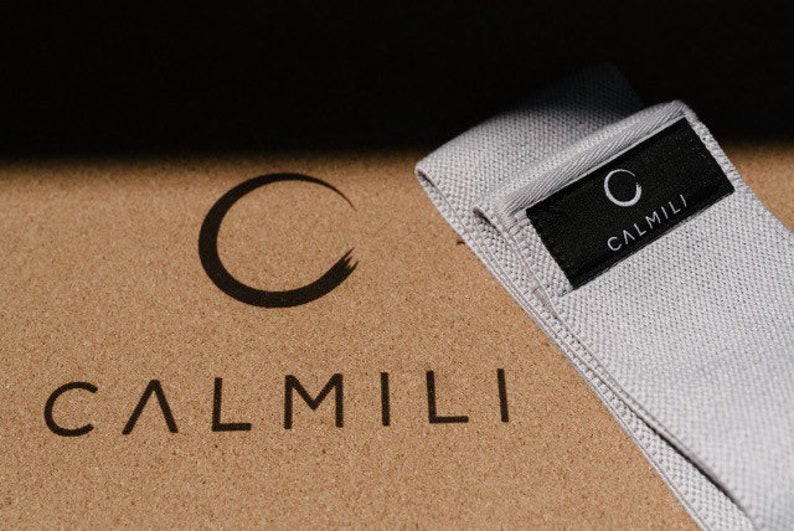 K&ouml;nnte beinhalten: Ein graues Stoff-Widerstandsband mit dem Wort "CALMILI" und einem Kreislogo, das auf einem schwarzen Etikett gedruckt ist. Das Band liegt auf einer Korkoberfl&auml;che mit dem Wort "CALMILI" in Schwarz gedruckt.