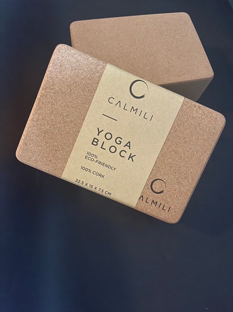 Puede incluir: Dos bloques de yoga rectangulares de corcho con bordes redondeados. Los bloques son de color marr&oacute;n claro natural. Uno de los bloques est&aacute; parcialmente cubierto por una etiqueta que dice "CALMILI YOGA BLOCK 100% ECO-FRIENDLY 100% CORK" y las dimensiones 22,5 x 15 x 7,5 cm.