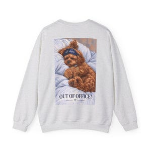 Peut inclure: Un sweat-shirt gris clair à col rond avec un motif rectangulaire au dos. L'image montre un chien brun aux poils bouclés dormant sur une literie à rayures bleues et blanches, portant un masque de nuit bleu marine avec l'inscription off duty.