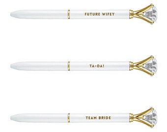 May include: Three white pens with gold accents and a diamond-shaped clear crystal on the end. The pens have the following text printed on them: "FUTURE WIFEY", "TA-DA!" and "TEAM BRIDE".