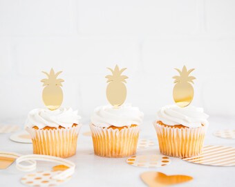 Puede incluir: Tres cupcakes blancos con adornos de piña dorados. Los cupcakes están sobre una superficie blanca con confeti dorado y blanco.