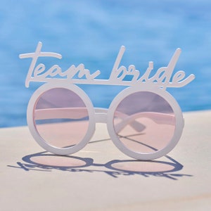May include: White plastic sunglasses with pink lenses and the words "Team Bride" written on the top.