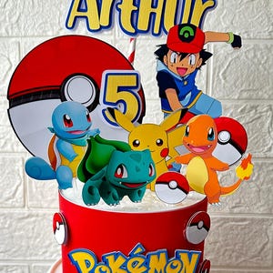 May include: A red cake decorated with Pokémon characters, including Pikachu, Charmander, and Bulbasaur. The cake features a topper with the name "Arthur" and the number "5". The Pokémon logo is displayed on the cake's side.