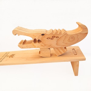 May include: A wooden alligator phone stand with a natural wood finish. The stand is made of wood and has a simple design. The alligator is standing on a wooden base with the words "GLACIER WOOD CRAFTS" engraved on the base.