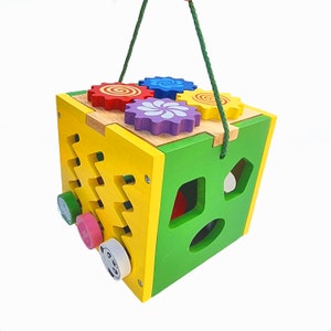 May include: A colorful wooden activity cube for toddlers. The cube features gears, shape sorters, and rotating pieces. The cube is painted in yellow, green, and natural wood tones, with red, blue, and purple gears. A green rope handle is attached.