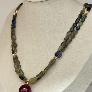 May include: A multi-strand necklace featuring small, polished, irregular pebble-shaped beads in shades of olive green, dark blue, and grey. The necklace is accented with small gold-toned spacer beads and a single, large, round, deep red pendant.