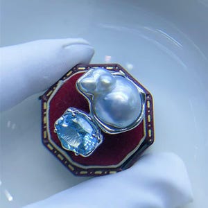 May include: A silver-toned jewelry piece featuring a large, lustrous, light-blue pearl and a smaller, oval-cut, light-blue gemstone. The gems are set on a red, octagonal base with a gold-colored trim. The background is a soft, neutral color.