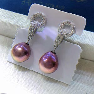 Pode incluir: Um par de brincos pendurados com pérolas roxas iridescentes em forma de pêra. Os brincos têm uma moldura prateada com um design circular brilhante no topo e um design linear que se conecta à pérola.