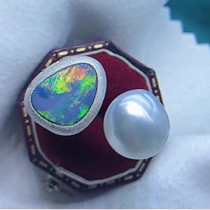 May include: A silver-toned ring with an oval, multi-colored opal stone next to a large, lustrous, gray pearl. The opal displays a vibrant array of colors, including blue, green, and orange. The jewelry is set against a red velvet background.
