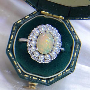 May include: A silver-toned ring featuring a central oval-shaped iridescent opal cabochon surrounded by a halo of small white pearls and a scalloped border of tiny clear stones, presented inside a dark green velvet-lined octagonal jewelry box.