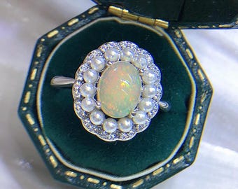 Könnte beinhalten: Ein silberfarbener Ring mit einem zentralen, ovalen, irisierenden Opal-Cabochon, umgeben von einem Kranz aus kleinen weißen Perlen und einem gewellten Rand aus winzigen klaren Steinen, präsentiert in einer dunkelgrünen, achteckigen Schmuckschatulle.
