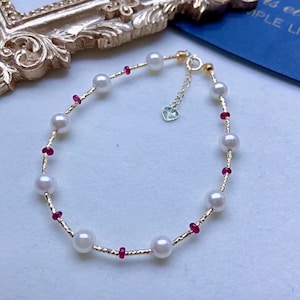 May include: Delicate gold bracelet featuring white pearls and small red gemstones. The bracelet has a delicate chain and a heart-shaped charm. A beautiful piece of jewelry.