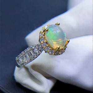May include: A gold and silver ring featuring a large, oval opal gemstone with a halo of small diamonds. The ring's band is encrusted with smaller, rectangular-cut diamonds. The opal displays a play of color, with hints of green and yellow.