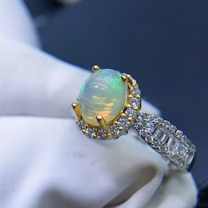 May include: A ring featuring a central oval cabochon opal with iridescent flashes of green and orange, set in yellow gold prongs. The halo and band are encrusted with numerous small, clear, round and baguette-cut white diamonds.