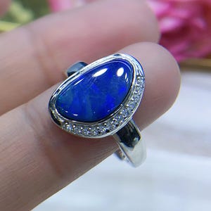 May include: A silver ring featuring a large, oval-shaped dark blue opal cabochon as the centerpiece. The opal is surrounded by a halo of small, clear, round-cut stones set into the silver band, which has a polished, reflective metallic finish.