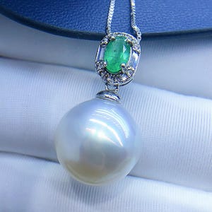 May include: A silver necklace featuring a large, lustrous white pearl pendant. The pendant is accented with a smaller, oval-cut green emerald surrounded by small diamonds. The necklace is displayed on a white fabric.