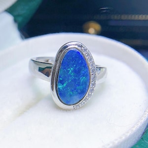 May include: A silver ring featuring a large, oval-shaped blue opal gemstone. The opal has a vibrant, iridescent quality. Small, clear gemstones are set along one side of the ring's band, adding sparkle. The ring is displayed on a white surface.