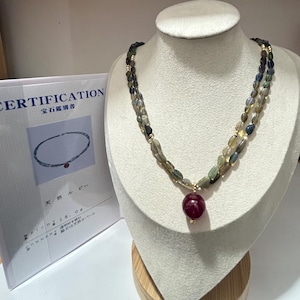 May include: A gemstone necklace displayed on a mannequin. The necklace features two strands of oval-shaped, multi-colored beads with gold accents. A large, round, deep red gemstone pendant hangs from the center. A certificate is visible in the background.
