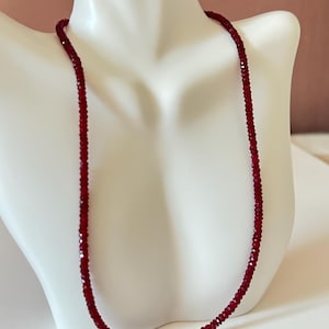 May include: A single strand necklace composed of small, deep red, faceted beads. The beads are uniform in size and shape, creating a continuous, circular line that rests against the neck form. The deep red color is consistent throughout the piece.