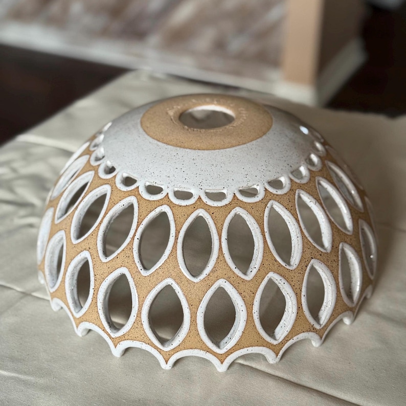 A handmade ceramic lamp shade in off-white and beige. The shade features a repeating pattern of cut-out teardrop shapes, creating a delicate, lacy effect. The top of the shade has a solid beige center, surrounded by a speckled off-white band. The shade is dome-shaped and has a scalloped edge. The overall style is bohemian and eclectic.