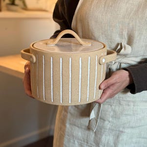 May include: A beige ceramic pot with a white striped design and a lid with a handle. The pot is being held by a person.