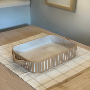 May include: A rectangular ceramic baking dish with white stripes around the rim and a handle on each side. The dish is sitting on a white and beige checkered tablecloth.
