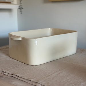 May include: A rectangular, off-white ceramic baking dish with rounded corners and two small handles. The dish sits on a neutral-colored, textured fabric, suggesting a kitchen or dining setting. The dish is empty and clean.