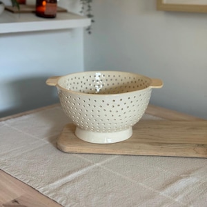 May include: Cream ceramic colander with many small holes. The bowl-shaped colander has a pedestal base and two small handles. It rests on a wooden board, suitable for kitchen use. The colander is likely used for draining food.