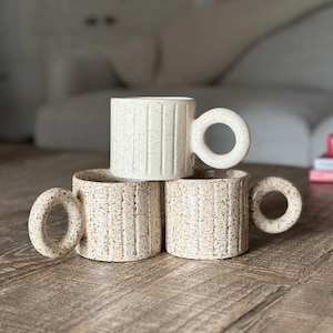 Op de afbeelding: Drie keramische mokken met gespikkelde witte glazuur en verticale lijnen. De mokken hebben grote, afgeronde handvatten.