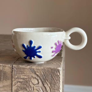 May include: A white ceramic mug with a unique handle and three colorful paint splatters in blue, purple, and pink.