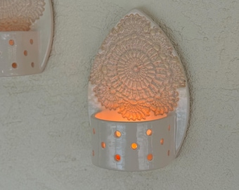 May include: A pair of off-white ceramic wall sconces, each featuring a teardrop-shaped backplate with an embossed lace doily pattern. The lower section of each sconce is a cylindrical holder with small circular cutouts that emit a warm glow.