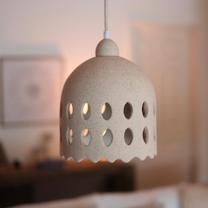 May include: A beige, speckled ceramic pendant light with a dome-shaped shade. The shade has evenly spaced oval cutouts and a scalloped bottom edge. The light hangs from a white cord and has a small, rounded top.