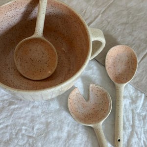 May include: A set of three speckled ceramic bowls in a light pink and white colour. The bowls are a large serving bowl, a smaller serving spoon, and a salad fork.