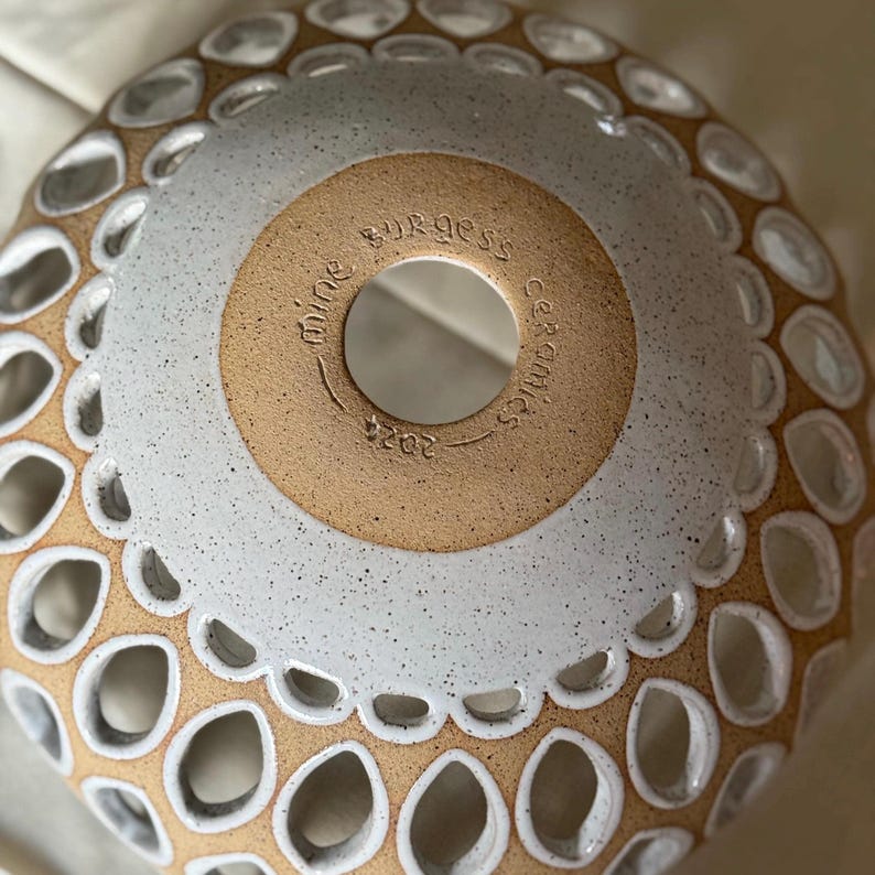 Close-up view of a ceramic bowl featuring a speckled off-white glaze and a tan-colored center band. The center band is stamped with the text 'Mine Burgess Ceramics 2022.' The bowl's edge is adorned with a repeating pattern of cut-out teardrop shapes, creating a decorative, lacy effect. The overall style suggests a handmade, artisanal aesthetic.