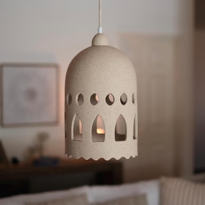 May include: Beige ceramic pendant light with a dome shape and a textured surface. The light fixture has arched cutouts and circular holes around the sides. A scalloped edge adorns the bottom. The light hangs from a beige cord.