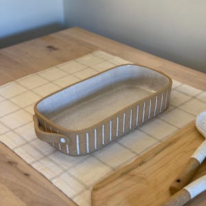 Puede incluir: Un plato para hornear rectangular de cerámica con rayas verticales blancas y un glaseado moteado. El plato tiene dos asas a los lados.