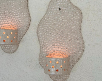 Op de afbeelding: Twee witte keramische wandlampen met een getextureerd oppervlak. Elke lamp heeft een ronde, opengewerkte kaarshouder. De kaarshouders zijn aangestoken en gloeien.