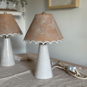 May include: Two ceramic table lamps with white bases and tan, textured shades. The shades have a scalloped edge. The lamps are on a neutral-coloured surface, with a power cord visible. The lamps are a decorative lighting fixture.