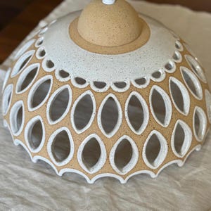 Two-tone stoneware dome shade resting on cloth; raw buff clay body transitions to white glazed crown, rows of leaf-shaped cutouts and scalloped edging—artisan pendant lamp detail shot.