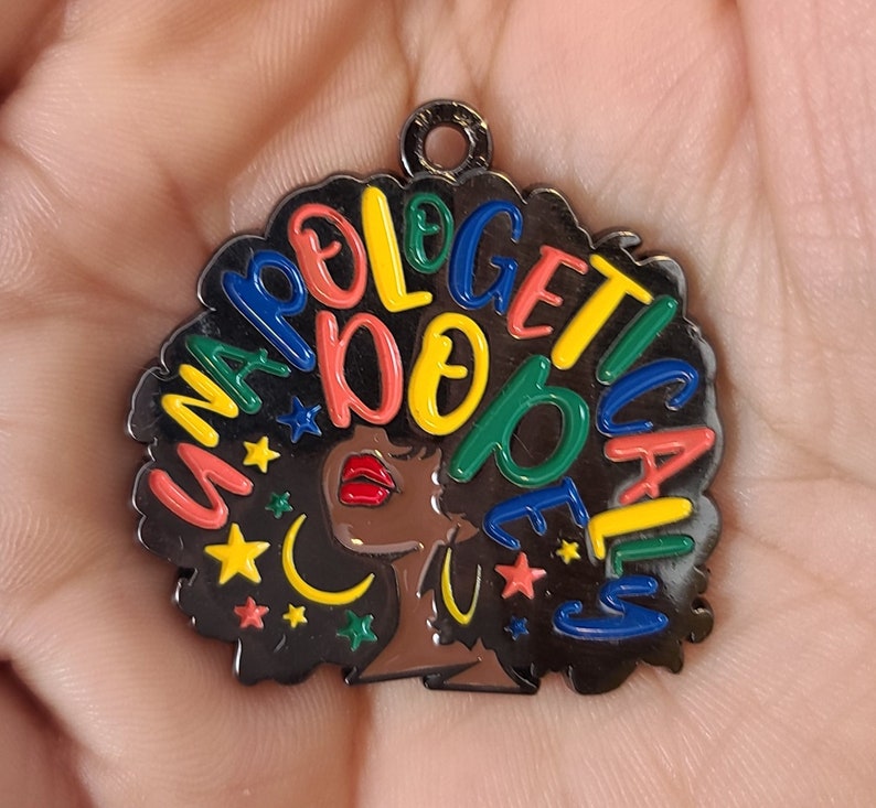 Op de afbeelding: Een zwarte emaille bedel met de woorden "Unapologetically Dope" in kleurrijke letters. De bedel toont een profiel van een vrouw met rode lippen en een zwarte afro, versierd met sterren en een maan.